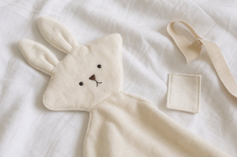 Doudou cousu main en forme de petit lapin en coton bio écru avec visage brodé, posé sur un drap blanc froissé doux
