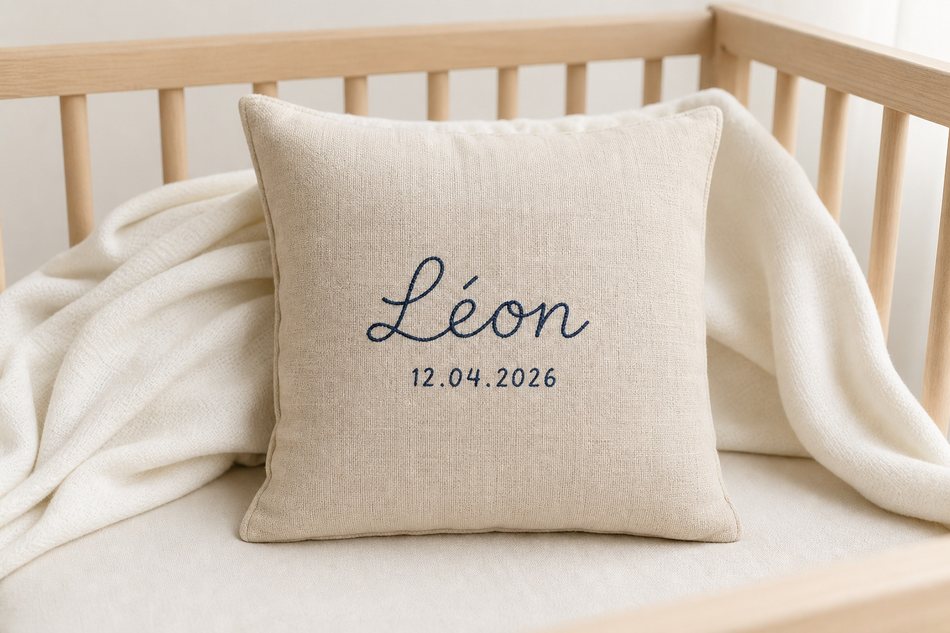 Coussin en lin écru 30×30 cm avec prénom brodé en script indigo sur lit de bébé minimaliste, lumière douce du matin