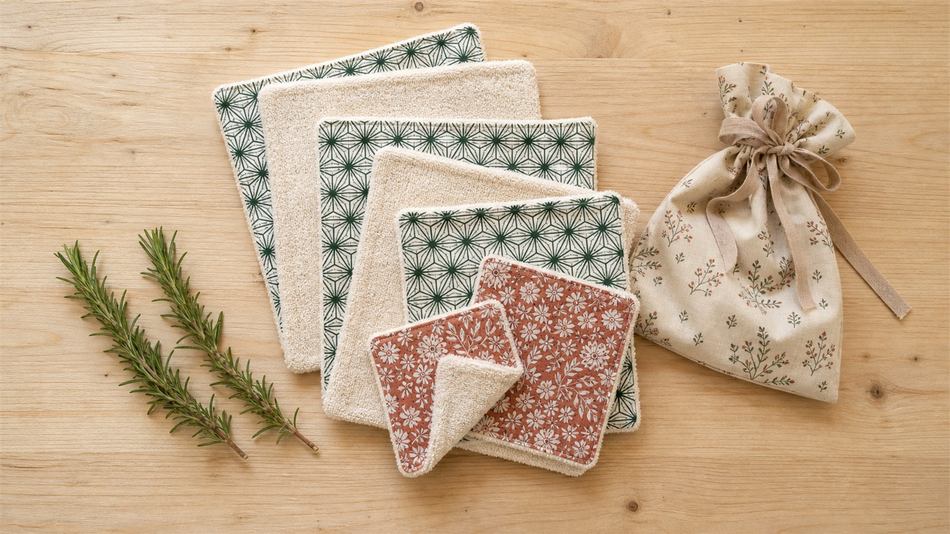 Set de 5 lavettes éponge écrues avec coton imprimé contrastant, pliées en pile à côté d'un sachet en tissu et d'un ruban beige