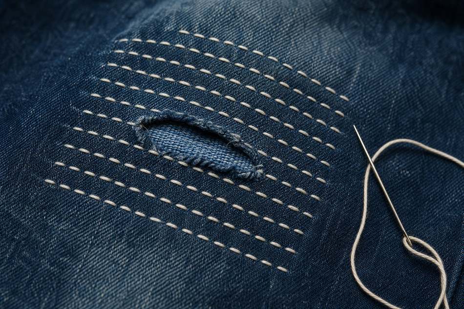 Détail d'un jean réparé avec du sashiko blanc sur le genou — lignes droites parallèles régulières avec tissu de renfort indigo apparent sous le trou